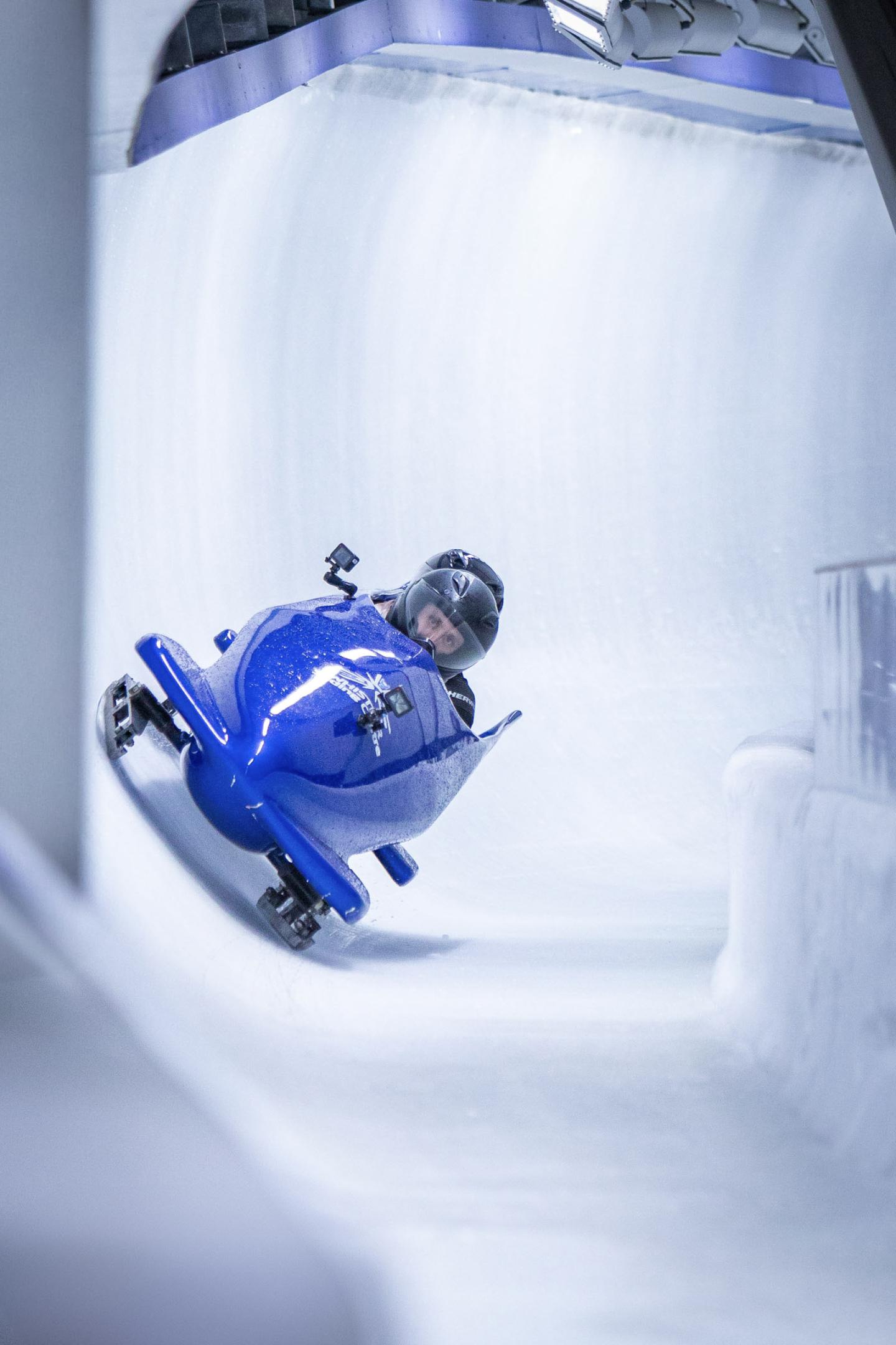 Blue bobsled speeding down icy track, driver focused and wearing a helmet.
